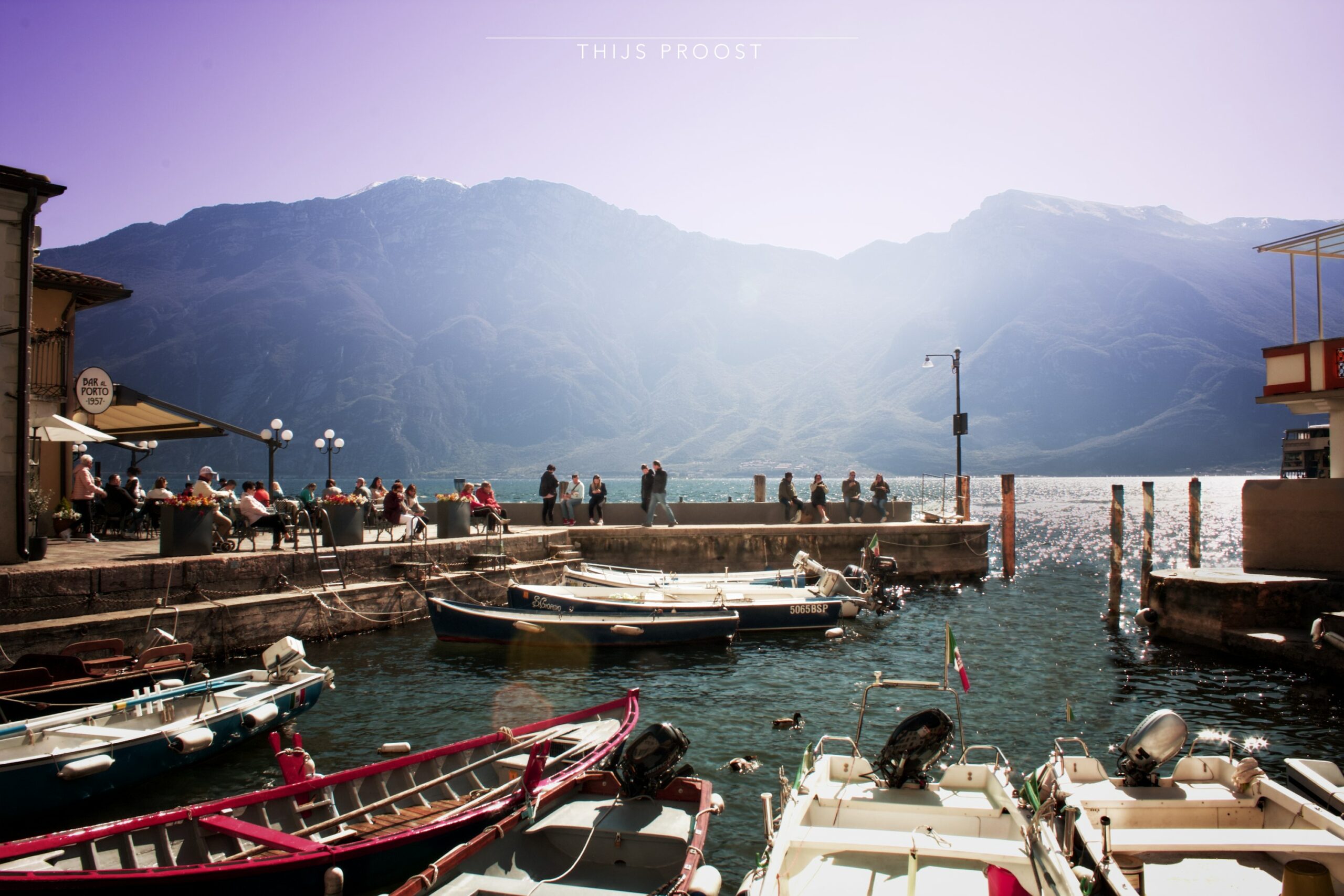 View of lake Garda. in the front are smal boots. A peer in front of the lake with people enjoying the sun. In the back are haze mountains with a clears blue sky.