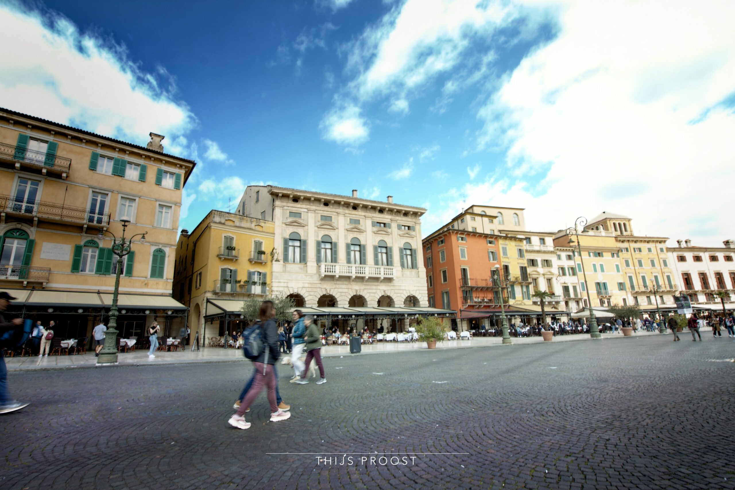 View of the market square of Verona Italy. In the background are buildings of classic Italian style. The from view shows people walking on their whey. They are a bit blurry because of motion blur. The sky is blue with medium clouds. it is a sunny day so the clouds are white.