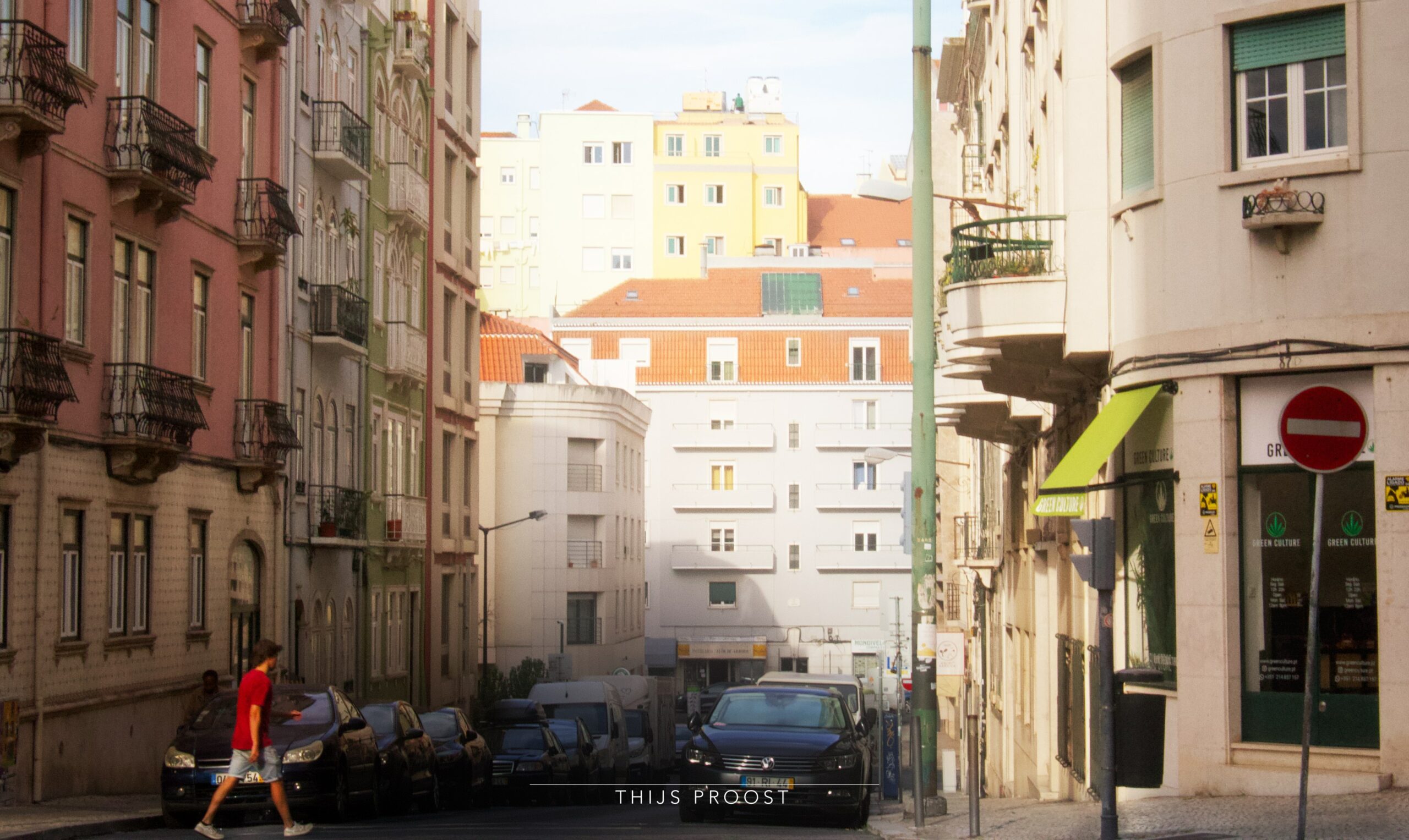 A man crosses a street in the center of Lisbon. Light false at the buildings at the end of the street. Cars are parked at the sides of the road.