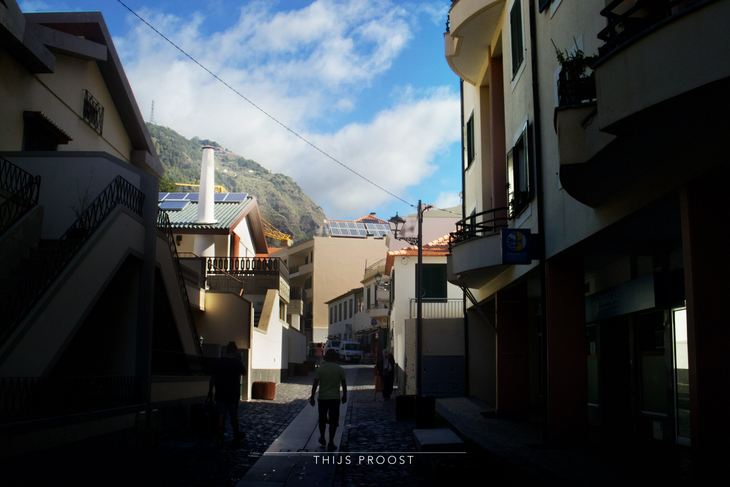 View af a street in the village of Ribeira Brava Madeira. The street is dark. The sun trows light in the tops of the building. In the distance you can see clouds in the sky. A mountain is precent at the background. People welk thought the street.