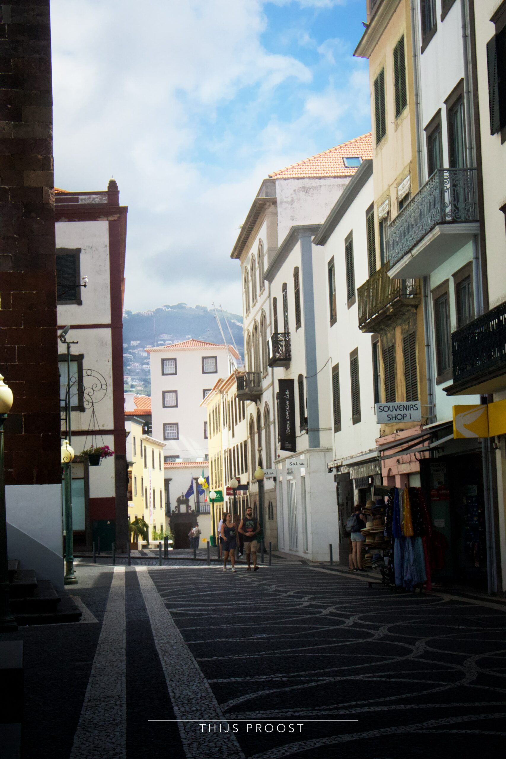 View of a street in Funchal Madeira. The houses on the right are in sunlight while the street remains in shadow. In the distance you see a hill and a cloudy sky. A the left you see a part of a church.