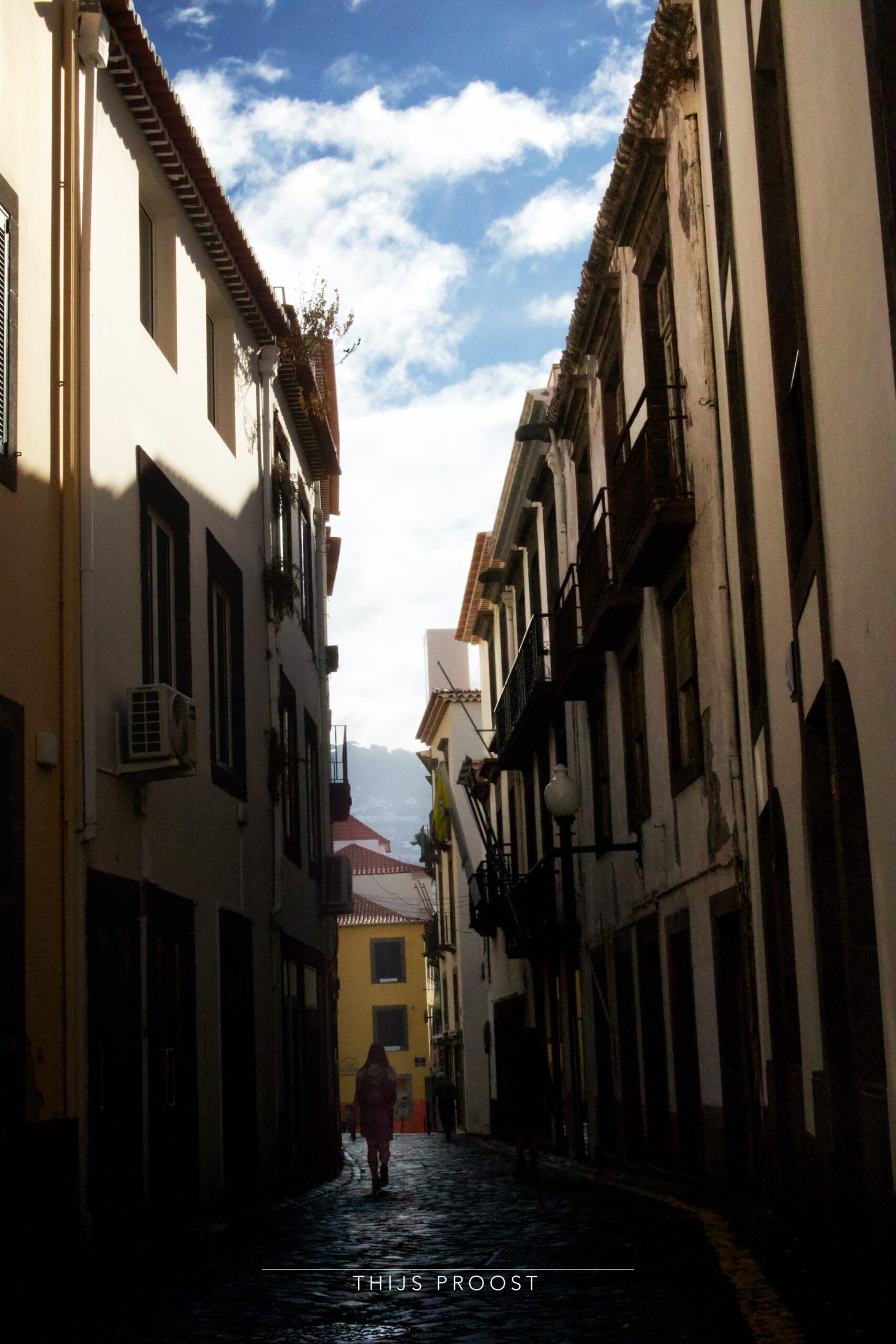 A view of a street in shadow in the Center of Funchal Madeira. A person walks in the dark. Sun hits the tips of the buildings. Clouds in the sky.
