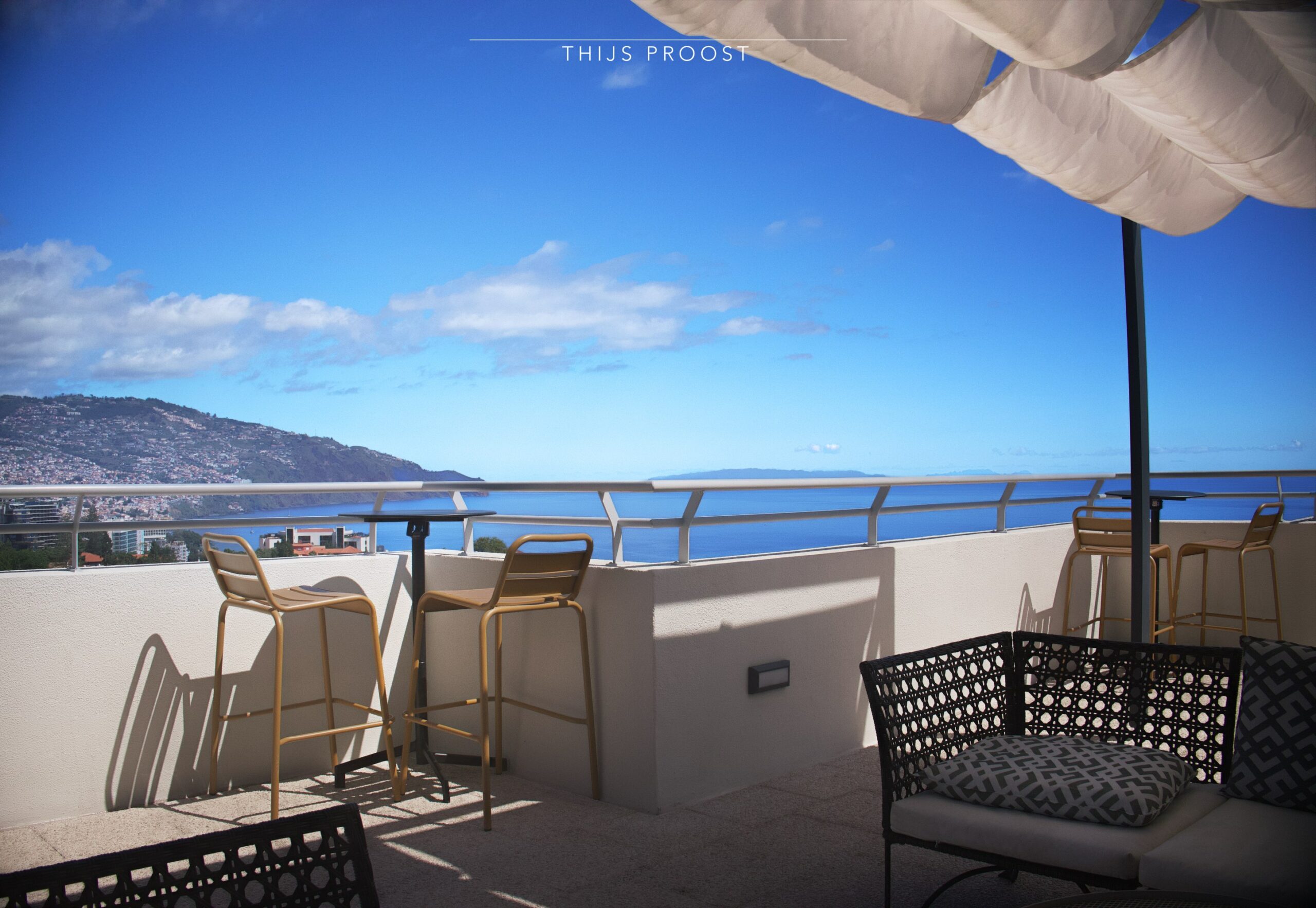 View on top of a roof terrace in Funchal Madeira. Seats are in the forgrond. The ocean in the background is blue as well ax the sky. At the left you can see part of the city of Funchal.